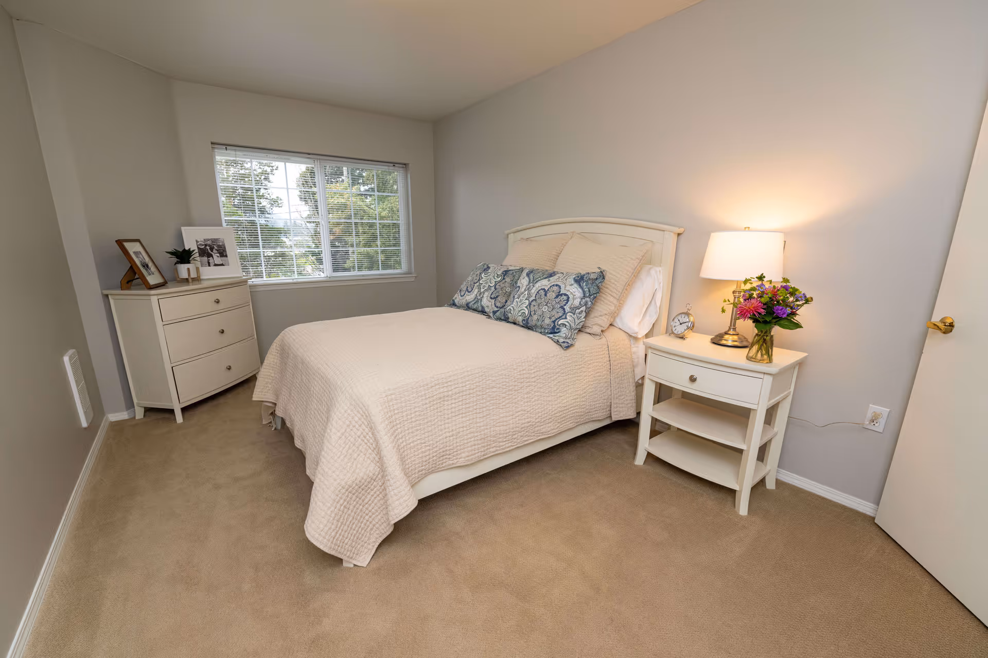 A neatly arranged bedroom with a beige carpet, a bed with a light beige quilt and decorative pillows, a white nightstand with a lamp, an alarm clock, and a vase of flowers, and a white dresser with framed photos and a small plant near a window with blinds.