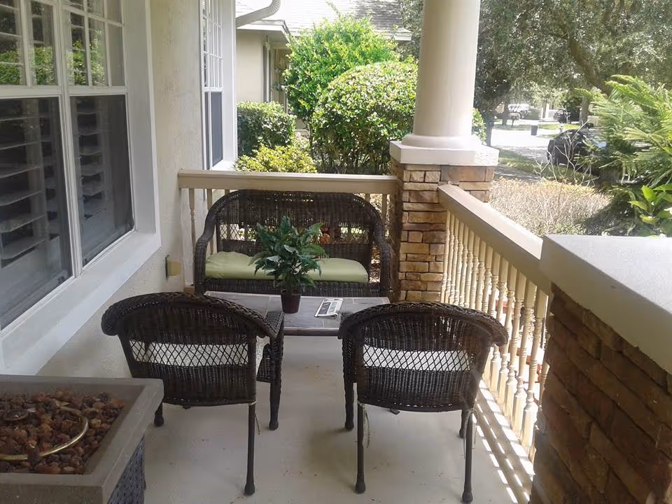 Front porch with wicker seating—two chairs and a bench—around a small table with a potted plant and railing overlooking the yard.