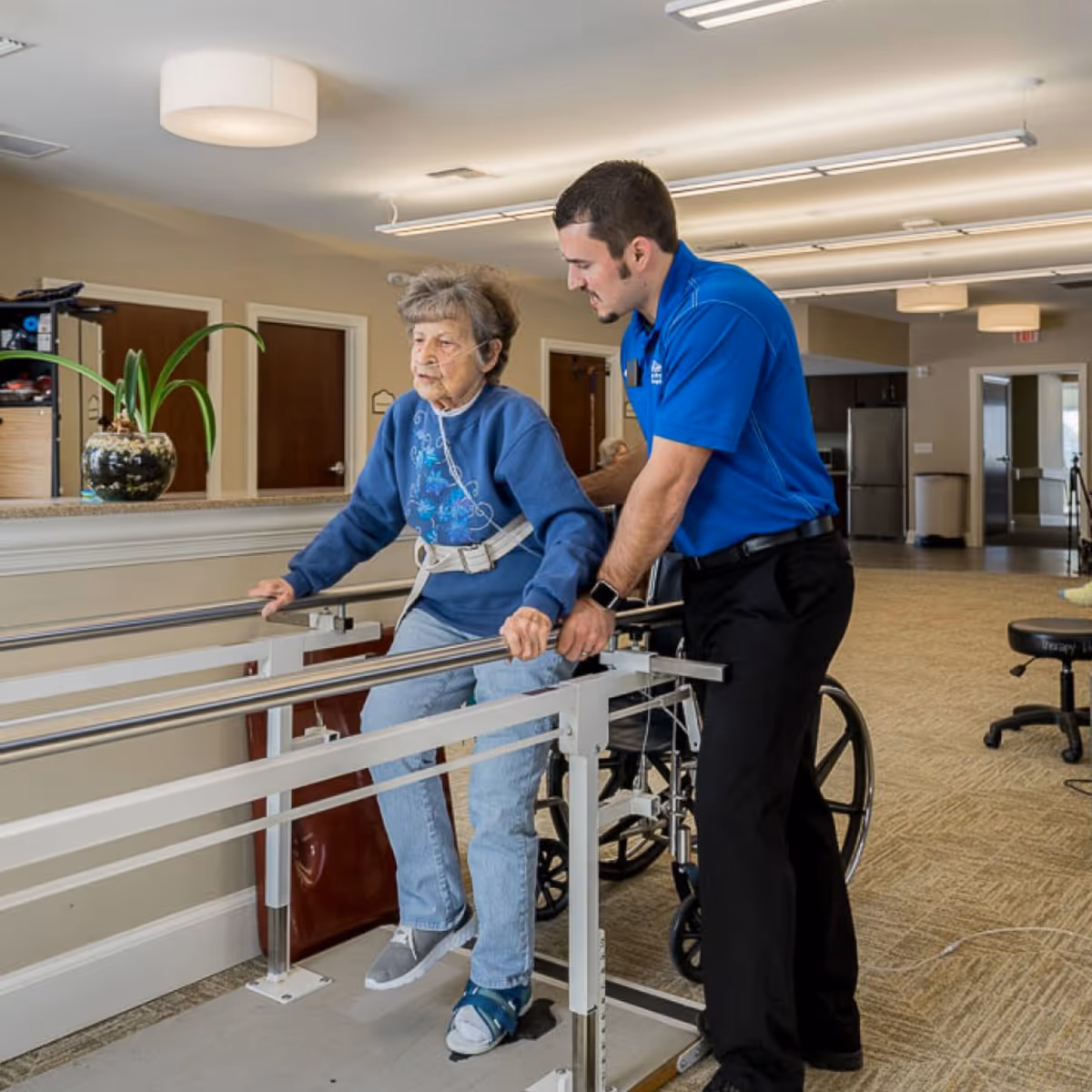 An elderly woman using parallel bars for physical therapy with the assistance of a male caregiver in a spacious, well-lit rehabilitation area. The woman is wearing a blue sweatshirt and jeans, and the caregiver is dressed in a blue polo shirt and black pants. A wheelchair is visible behind them, and the room has beige walls and carpeted floors.