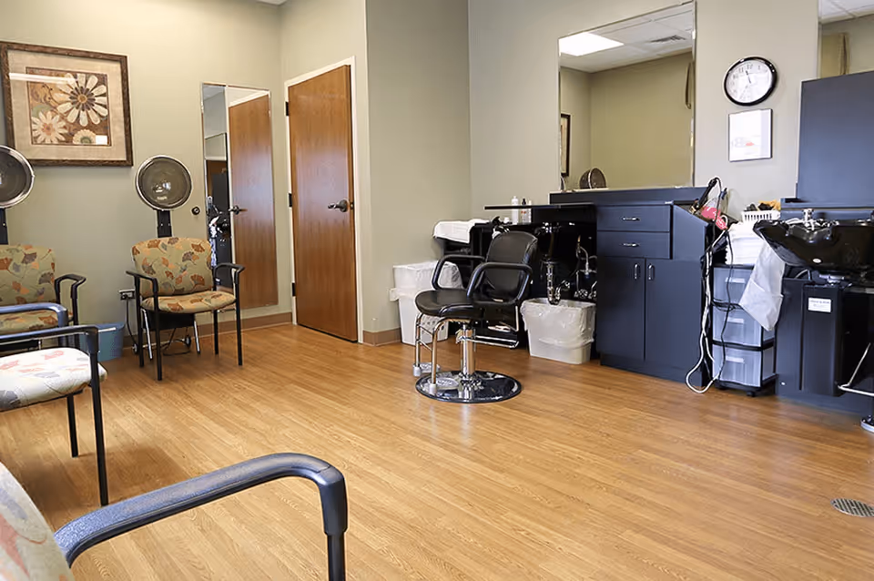 Interior view of a hair salon area in a senior living facility with wooden flooring, salon chairs, hair washing stations, a large mirror, and a few patterned waiting chairs. A clock and framed artwork are visible on the walls.