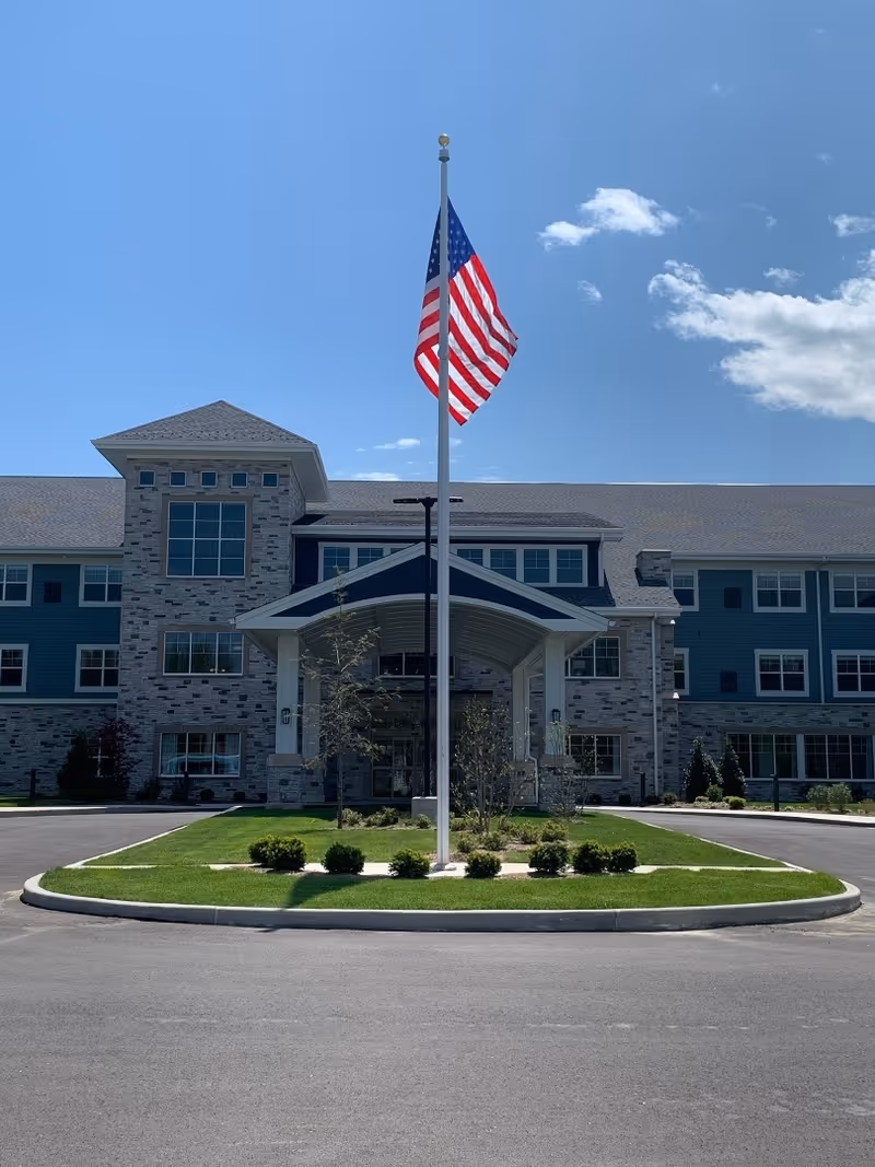 Front exterior view of a large senior living facility building with a covered entrance and an American flag on a flagpole in the center of a circular driveway. The building has multiple windows and a combination of stone and siding on the facade under a clear blue sky.