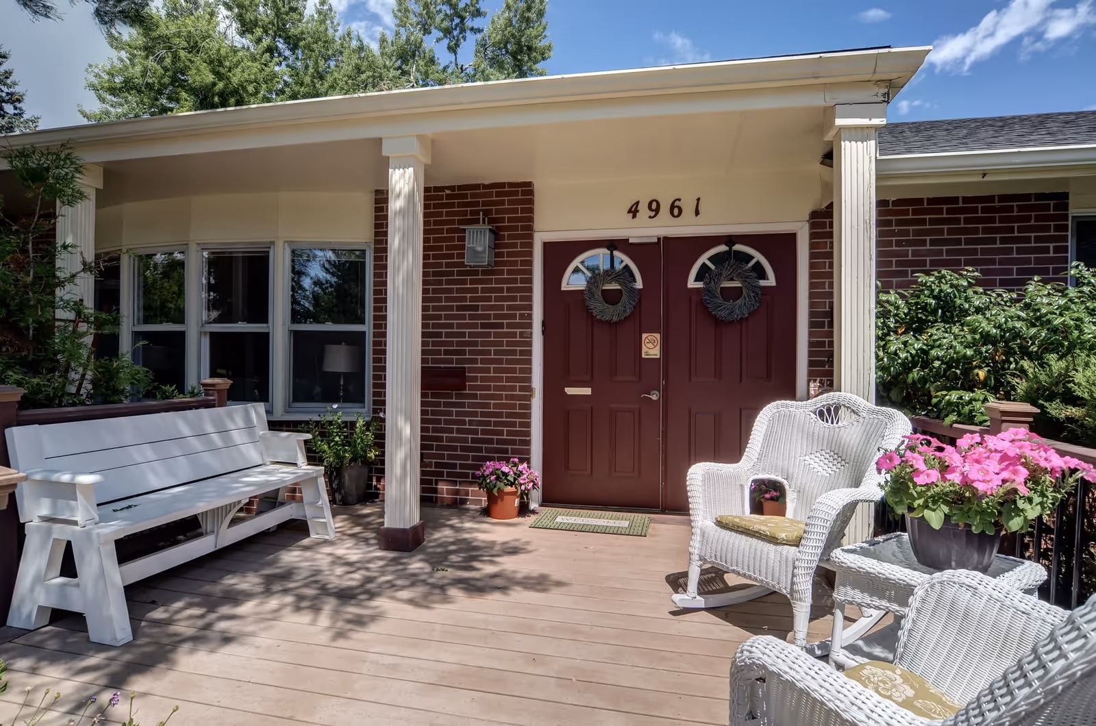 Front porch of a brick building with the address number 4961 above double maroon doors decorated with wreaths. The porch has white wooden benches and wicker chairs with cushions, a small table with a pot of pink flowers, and some potted plants near the entrance. Trees and bushes surround the porch area under a clear blue sky.