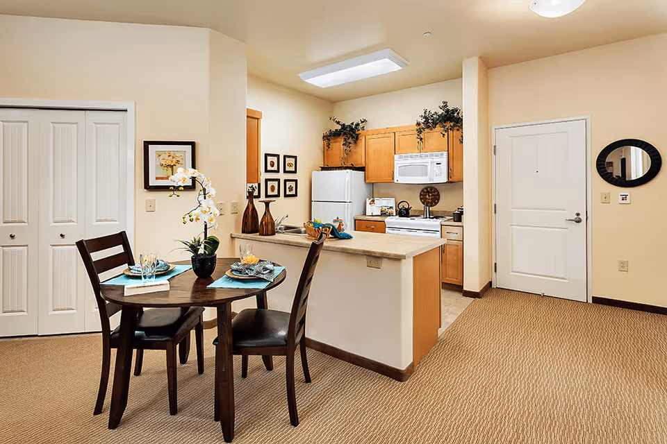 Round dining table with place settings and chairs next to an open kitchen with light wood cabinets and white appliances in a warm-toned apartment.
