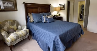A bedroom with a large wooden headboard bed covered with a blue quilt and multiple pillows. To the left of the bed is a patterned armchair with a hat on it. A nightstand with a lamp and framed pictures is next to the bed. An open doorway leads to a bathroom in the background.