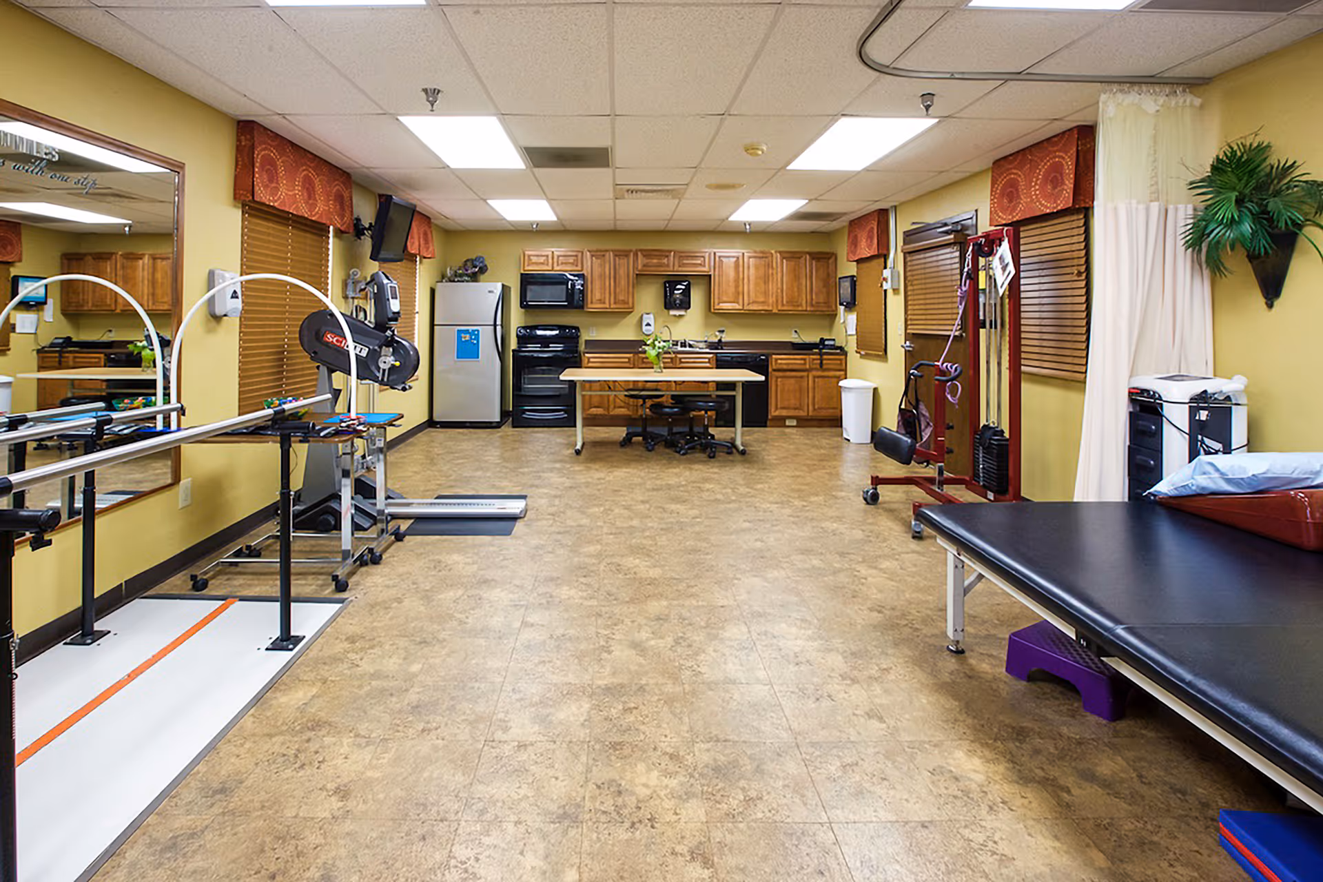A spacious rehabilitation therapy room with parallel bars, exercise equipment, a treatment table, and a kitchenette along the back wall.