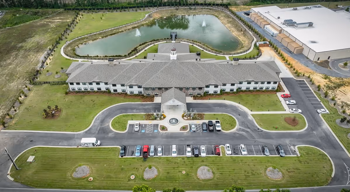Aerial view of The Fountains of Calhoun senior living facility showing a large two-story building with a gray roof, surrounded by well-maintained green lawns and a curved driveway with parked cars. Behind the building is a pond with two water fountains and a walking path around it. There is also a large warehouse-type building visible to the right.