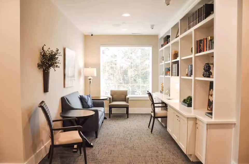 A cozy living area with a large window letting in natural light. The room features a blue sofa with two cushions, two armchairs, a round side table, a floor lamp, and a built-in white bookshelf filled with books and decorative items. The walls are light-colored, and the floor is carpeted.