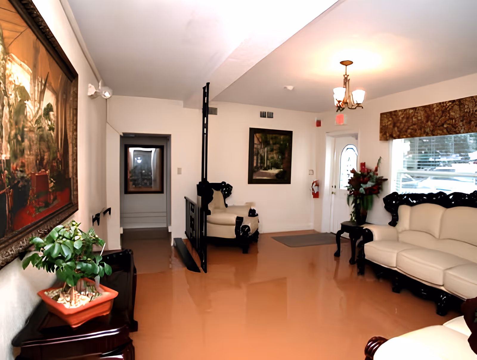 Bright senior living lounge with cream sofas and dark wood trim, a chandelier, framed paintings, and a potted plant near the entrance.
