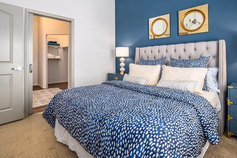 A bedroom with a large bed featuring a tufted headboard and blue and white patterned bedding. There are multiple pillows on the bed. On the right side, there is a blue nightstand with a lamp. Two round mirrors hang on the blue accent wall above the bed. An open door reveals a walk-in closet with shelves and folded towels.