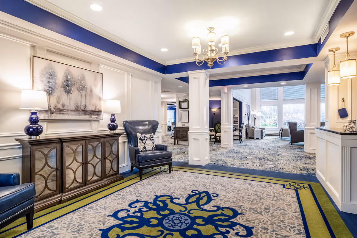 A spacious and elegant interior common area with white paneled walls and blue accents. The room features a large decorative carpet with blue and green patterns, two blue leather armchairs with patterned cushions, a wooden cabinet with two blue lamps, and a chandelier hanging from the ceiling. In the background, there are more seating areas with large windows letting in natural light.