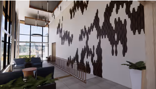 Modern interior lounge area with large windows allowing natural light, featuring dark gray sofas, small round tables, a tall white planter with greenery, and a decorative wall installation with dark hexagonal shapes arranged in a wave pattern.