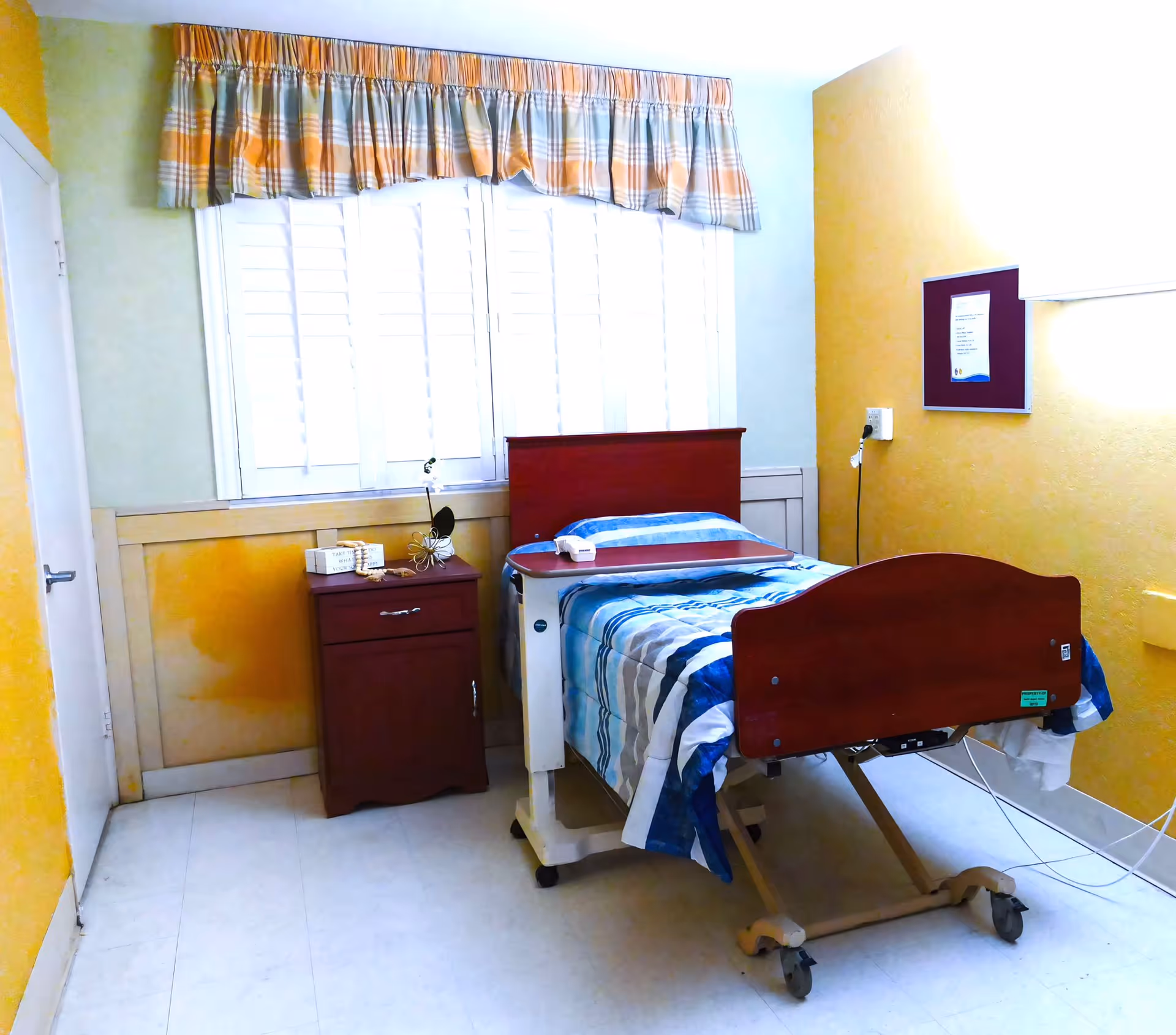 A small, simple patient room with a single hospital bed covered in a blue and white striped blanket. The bed has a wooden headboard and footboard and is equipped with wheels. Next to the bed is a wooden nightstand with a few items on top, including a small decorative piece and a box. The room has yellow walls, a window with white shutters, and a plaid valance curtain in shades of orange and gray. A bulletin board is mounted on the wall to the right of the bed.