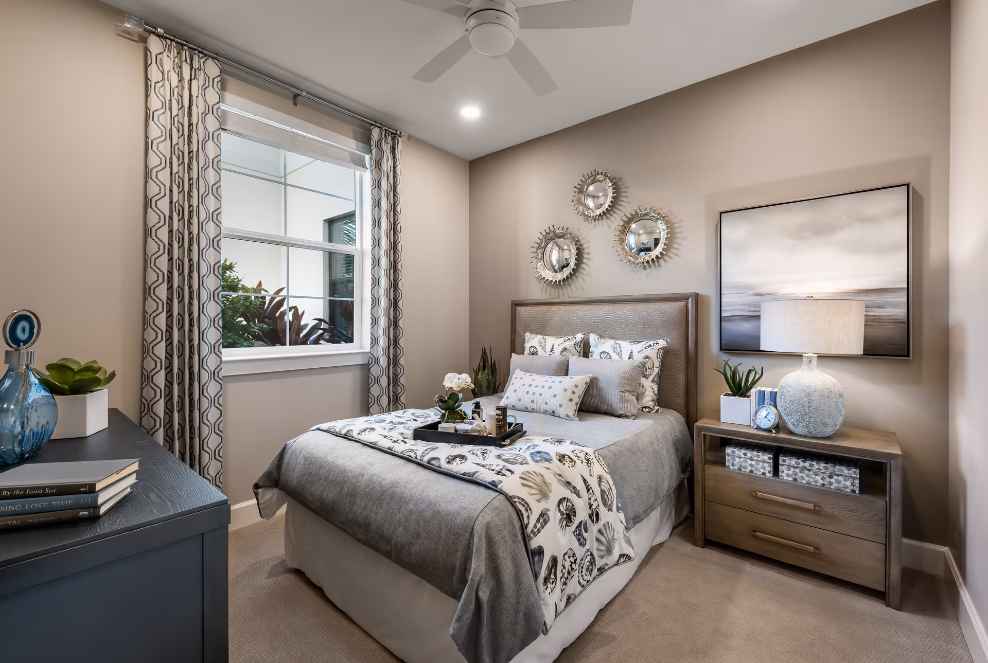 A cozy bedroom with a neatly made bed featuring gray and patterned bedding. Above the bed are three decorative round mirrors. To the right of the bed is a wooden nightstand with a lamp, a small plant, books, and a clock. To the left is a dark dresser with books and decorative items. A window with patterned curtains lets in natural light, and a ceiling fan is mounted on the ceiling.