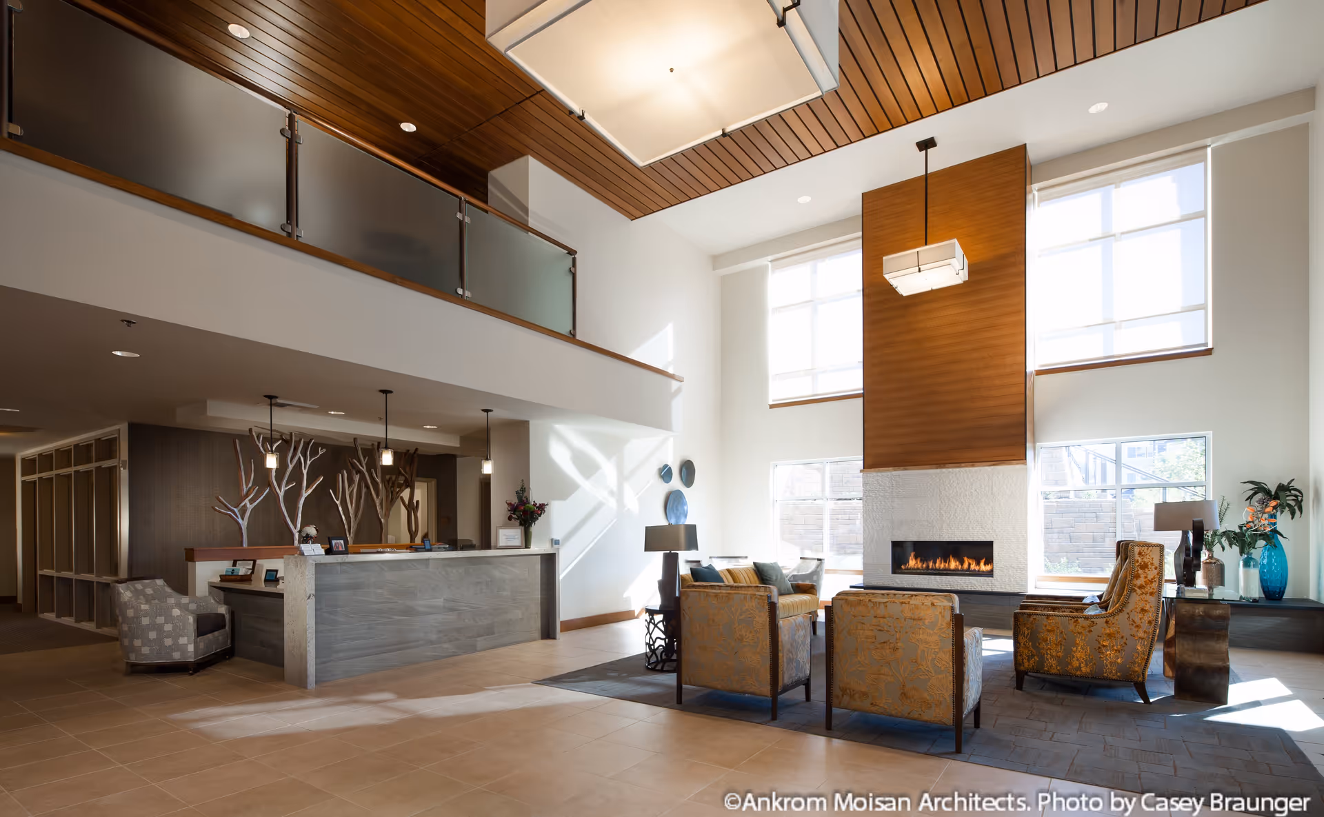 A spacious and modern senior living facility lobby with a high ceiling and large windows allowing natural light to fill the room. The area features a seating arrangement with four upholstered chairs around a rectangular fireplace with a wooden accent wall above it. To the left is a reception desk with pendant lights hanging above and decorative elements behind it. The ceiling has wooden paneling and a large square light fixture.