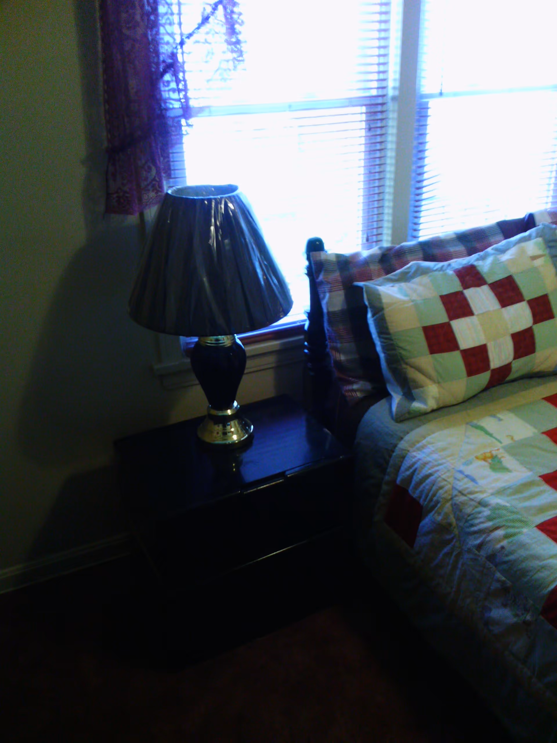 A bedroom corner with a wooden nightstand holding a lamp with a dark base and a shiny lampshade. Next to the nightstand is a bed with a wooden headboard, covered with a patchwork quilt and matching pillows in red, white, and green colors. Behind the nightstand and bed are two windows with blinds partially open and a purple curtain on the left window.
