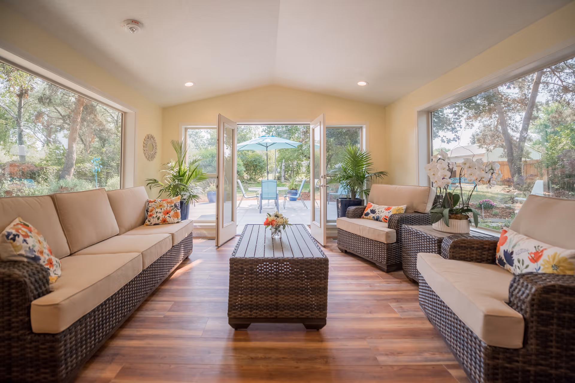 Sunlit living area with wicker sofas, a central coffee table, large windows and open doors leading to a patio.