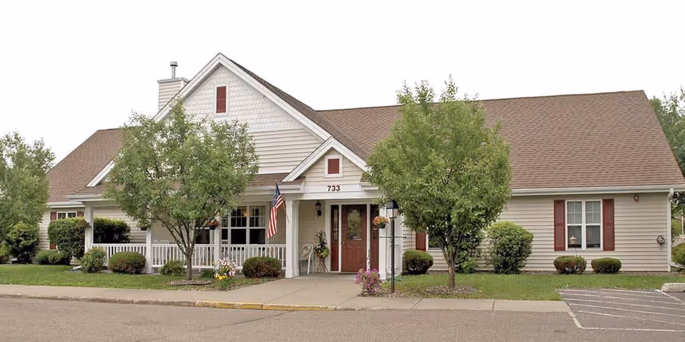 Exterior front view of a single-story senior living facility building with beige siding, a brown roof, and a covered porch. There are two trees and several bushes in the landscaped front yard, an American flag hanging near the entrance, and a paved driveway and parking area in front.