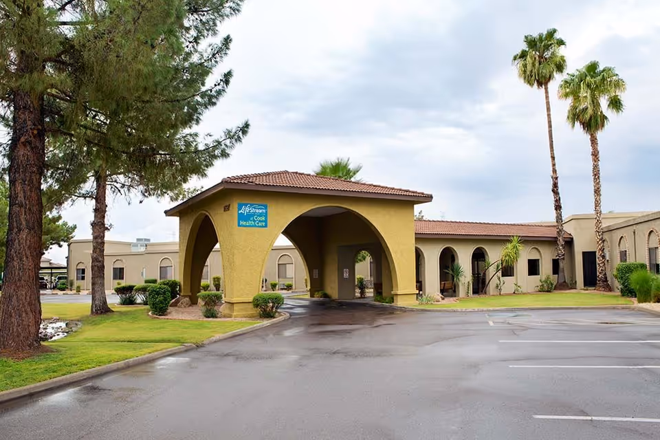 Drive-through arched entrance of a single-story Lifestream senior living building with palm trees and a wet parking lot.