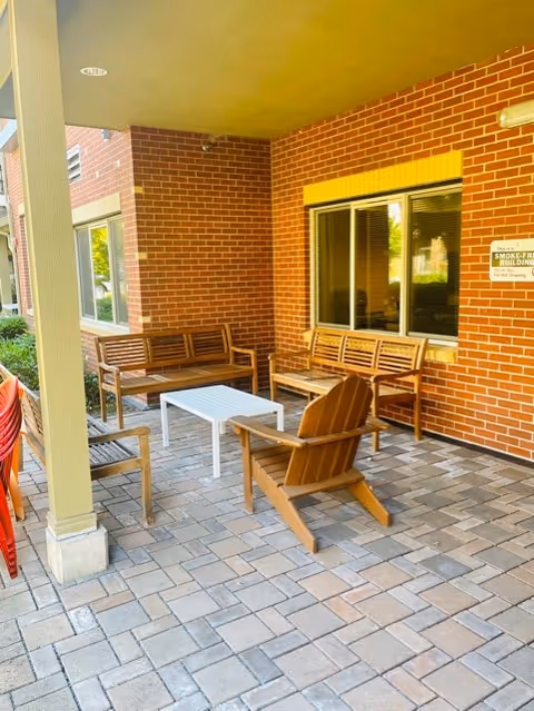 Covered outdoor patio area with brick walls, featuring wooden benches, a wooden chair, and a white rectangular table on a paved stone floor. There is a window with a yellow frame and a sign indicating the building is smoke-free.