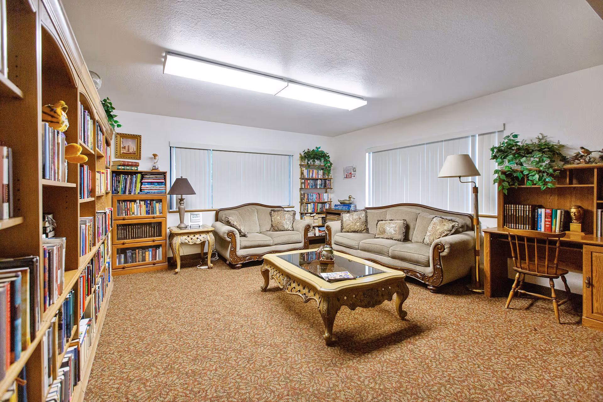 A cozy living room with two beige sofas, a glass-top coffee table with ornate legs, and a wooden side table with a lamp. The room has carpeted flooring and two large windows with vertical blinds. There are multiple bookshelves filled with books and decorative plants, as well as a wooden desk with a chair and a lamp on the right side.