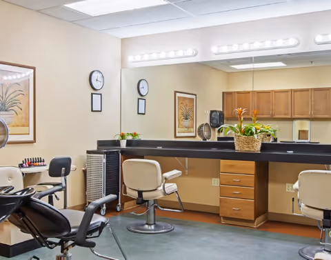 Interior of a salon area in a senior living facility with salon chairs, a large mirror with bright vanity lights, a countertop with a basket of plants, wall clocks, framed artwork, and cabinets in the background.