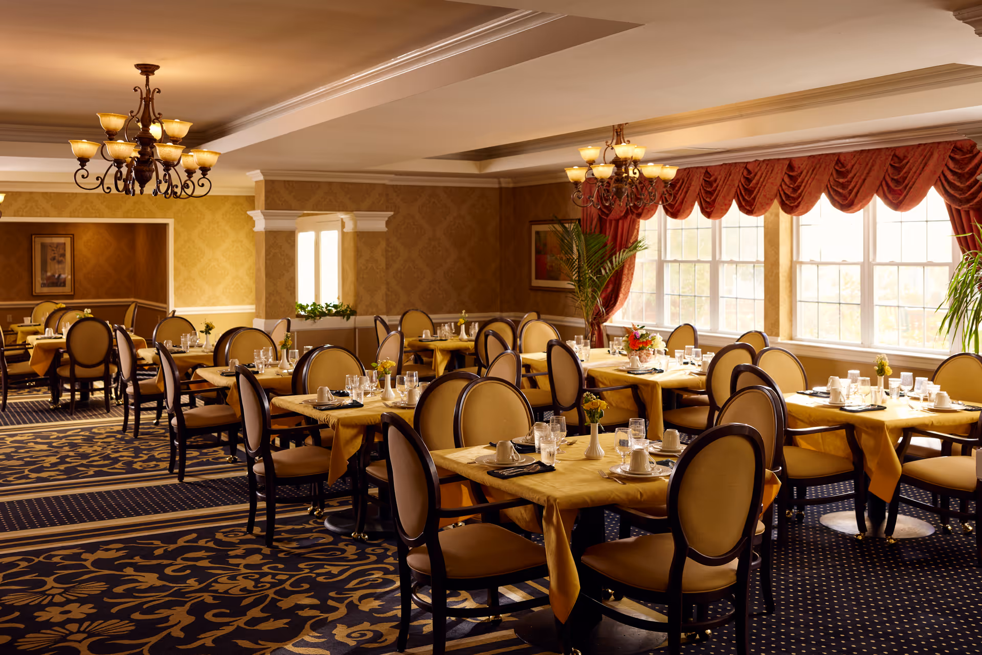 A warmly lit dining room with multiple tables covered in yellow tablecloths, each set with glasses, cups, napkins, and small flower vases. The room features patterned carpet, elegant chandeliers, large windows with red curtains, and classic-style chairs with cushioned seats and backs.