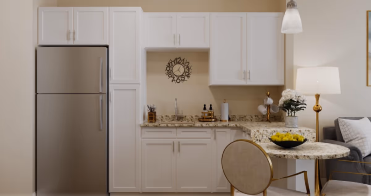 A modern kitchen area with white cabinets, a stainless steel refrigerator, a granite countertop with a sink, and a round granite table with a bowl of lemons. There is a decorative clock on the wall, a lamp with a white shade, a flower pot with white flowers, and a gray sofa with pillows partially visible.