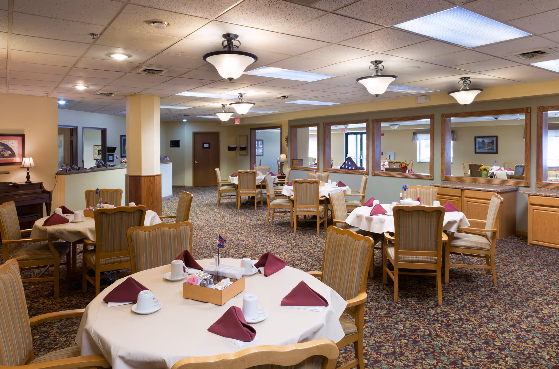A dining room in a retirement community with round tables covered in white tablecloths, each set with cups, saucers, and folded burgundy napkins. The room has patterned carpet, wooden chairs with cushions, large mirrors on the walls, and ceiling lights providing warm illumination.