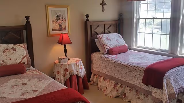 Sunlit bedroom with two twin beds dressed in floral quilts, a small round bedside table with a lamp, and a window.