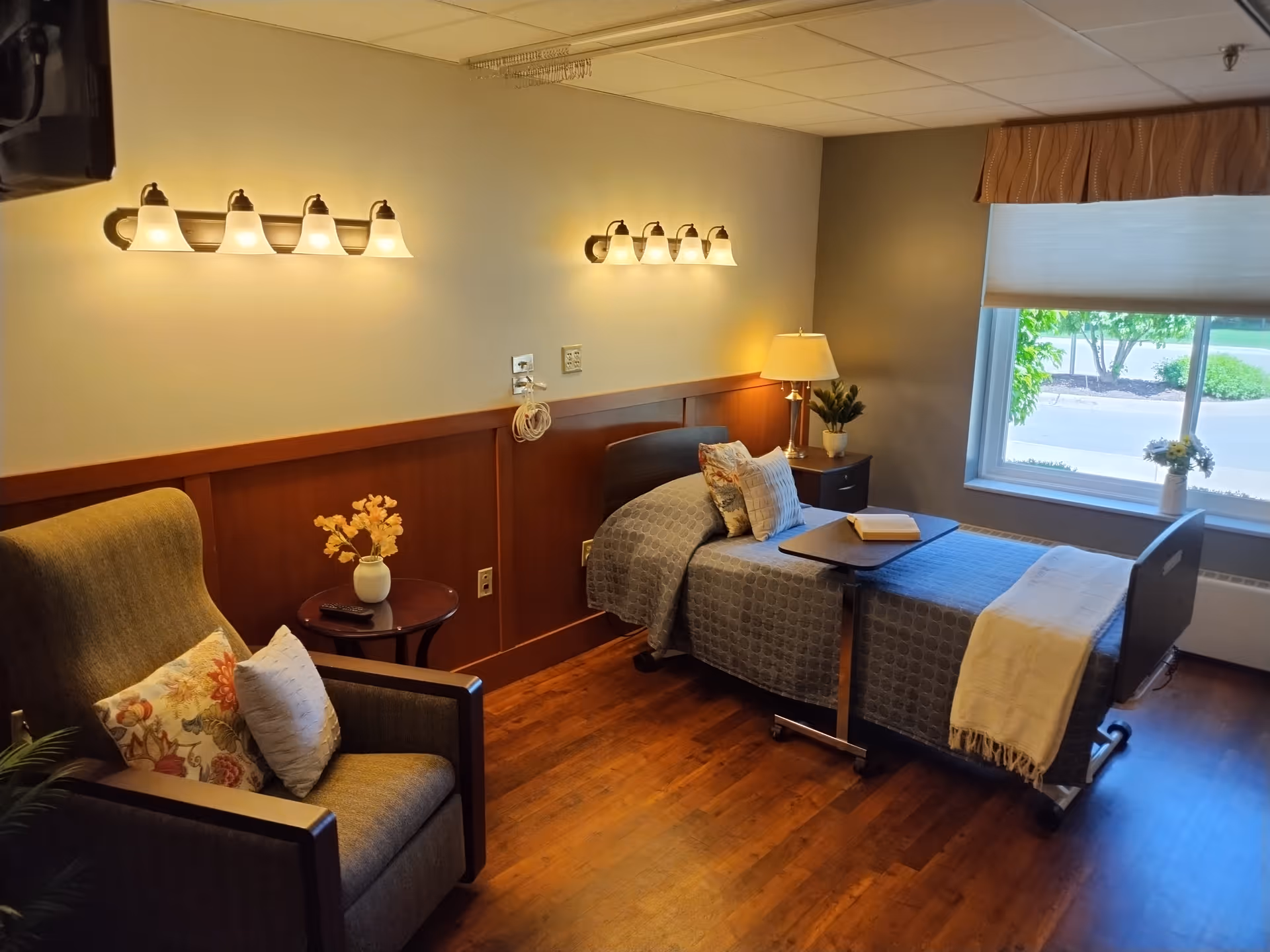 A cozy senior living bedroom with a single bed covered in a gray patterned blanket and a cream throw. The bed has pillows and a small overbed table with a book on it. Next to the bed is a nightstand with a lamp and a small plant. There is a large window with a valance and a view of greenery outside. A comfortable armchair with floral and white pillows sits beside a small round table with a vase of flowers. The room has warm wood flooring and soft lighting from wall-mounted fixtures.