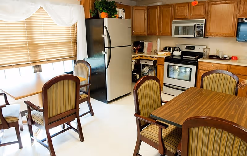Communal kitchen and dining area with wooden cabinets, a stainless refrigerator and stove, and a table surrounded by upholstered chairs.