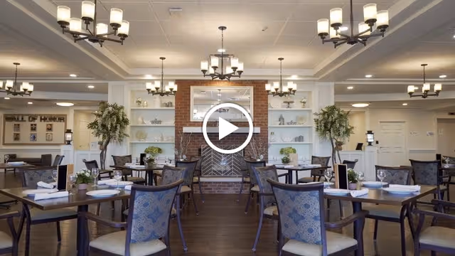 A spacious and well-lit dining room in a senior living facility featuring multiple tables set with white napkins and menus. The room has elegant chandeliers hanging from the ceiling, a brick fireplace with a mirror above it, built-in white shelves with decorative items, and potted plants on either side of the fireplace. The overall decor is warm and inviting.