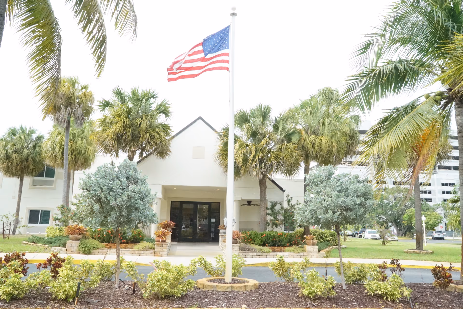 Entrance of a senior living facility with an American flag on a central flagpole, palm trees, and landscaped grounds.