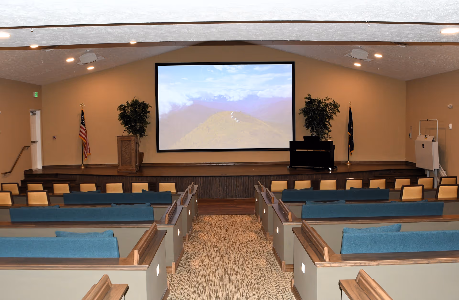 Auditorium-style room with rows of pew-like seating facing a stage with a large projection screen, piano, podium, and flags.