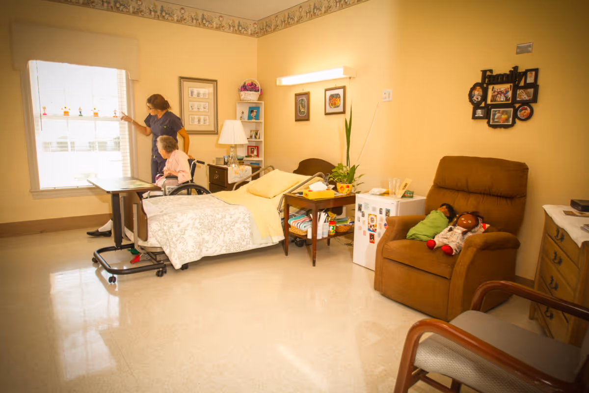 A nursing home room with a bed, a bedside table, a brown recliner chair with two dolls on it, a small refrigerator, and a nurse assisting an elderly woman in a wheelchair near a window with blinds. The walls are decorated with framed pictures and a family photo collage.