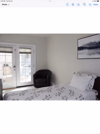 A bedroom with a single bed covered in white bedding with a black floral pattern. There is a black armchair in the corner next to a set of French doors with blinds. A framed picture of a mountain landscape hangs on the wall above the bed.