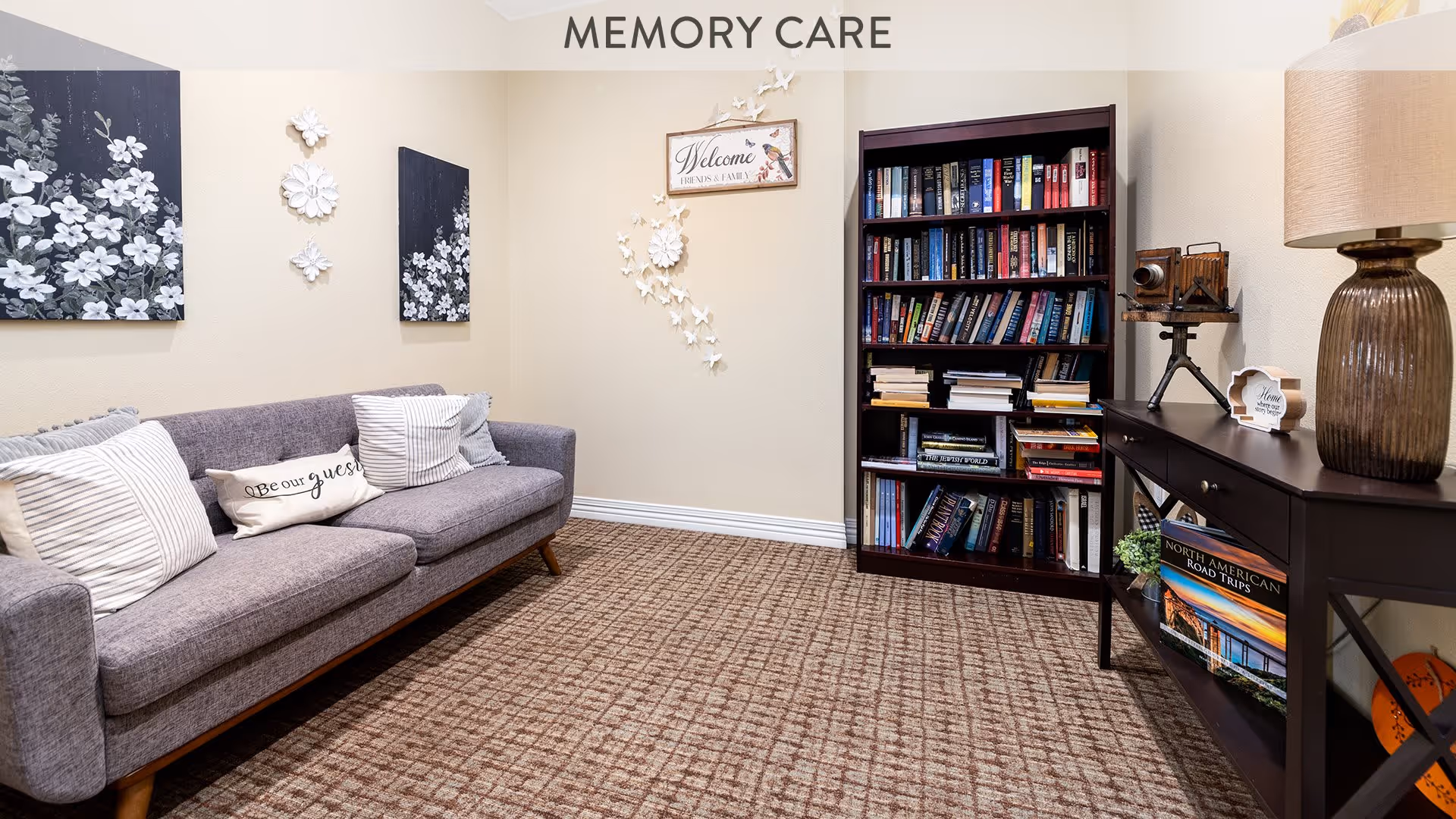 A cozy memory care room with a gray sofa adorned with striped and decorative pillows, a bookshelf filled with books, a dark wooden console table with a lamp and decorative items, and floral wall art on beige walls.