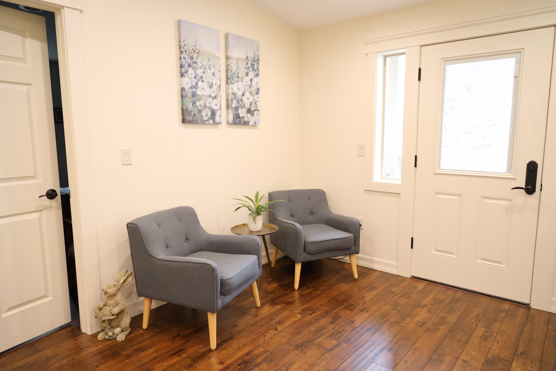 A small seating area near the entrance door featuring two gray armchairs with wooden legs, a small round side table with a potted plant, floral artwork on the wall, and a decorative rabbit figurine on the floor next to a closed white door.