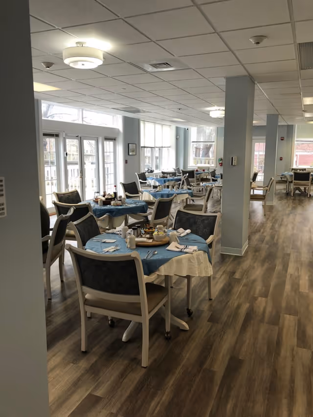 A bright dining room with multiple round tables covered with blue and white tablecloths, each set with napkins, utensils, and condiments. The room has large windows letting in natural light, light blue walls, and wood-patterned flooring. Several chairs are arranged around the tables.