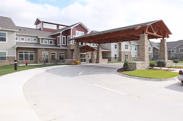 Front exterior view of Oak Park Place Dubuque building with a covered entrance supported by stone pillars, a curved driveway, and landscaped greenery under a partly cloudy sky.