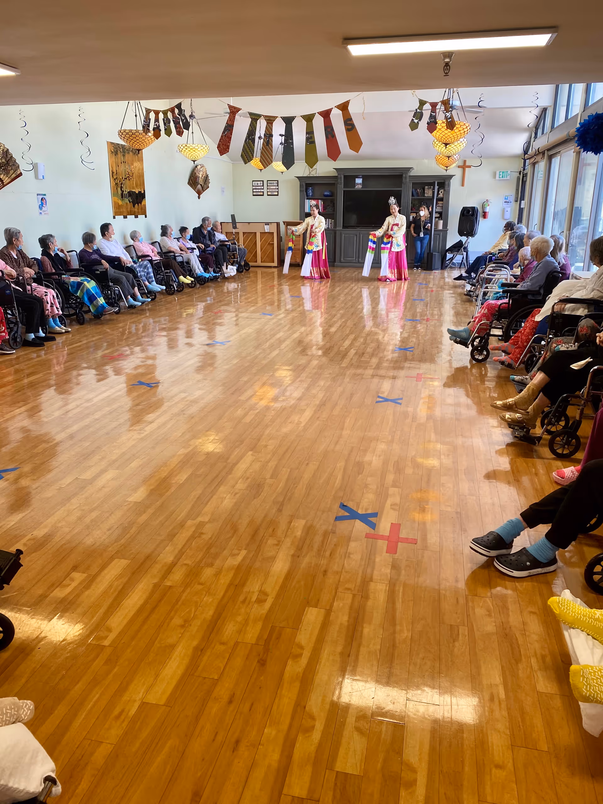 A large room with wooden floors and two performers in traditional attire dancing in the center. Elderly people are seated in wheelchairs along both sides of the room, watching the performance. The ceiling is decorated with hanging ties and lights, and there is a piano and a large cabinet with a TV at the far end of the room.