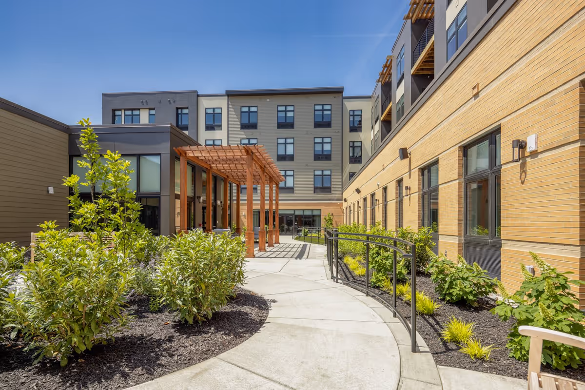 Outdoor courtyard area of a senior living facility with a curved concrete walkway, green shrubs and plants on both sides, a wooden pergola structure, and multi-story building facades with large windows under a clear blue sky.