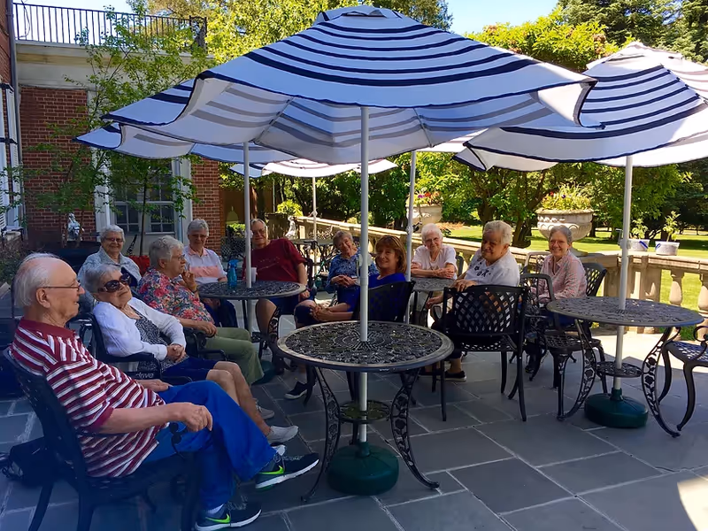 A group of elderly people sitting outdoors around metal tables with large white and blue striped umbrellas, enjoying a sunny day on a stone patio with greenery and a brick building in the background.