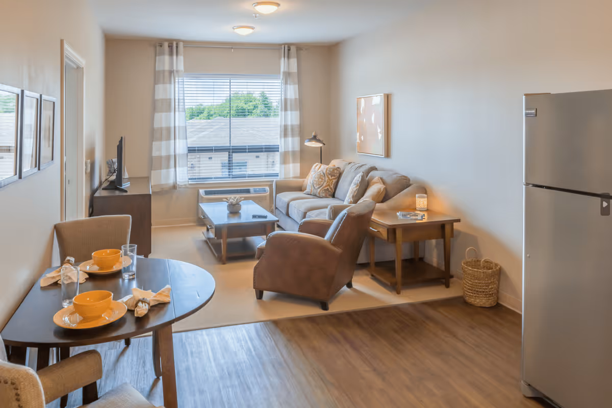 A cozy senior living room and dining area with a beige sofa, brown armchair, wooden coffee table, and side table with a lamp. The dining area has a round table set with two place settings including yellow bowls and glasses. A large window with striped curtains lets in natural light, and a stainless steel refrigerator is visible on the right side.