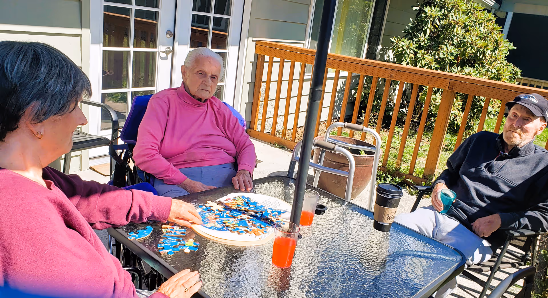 Three elderly individuals sitting around a glass outdoor table on a patio. Two women and one man are engaged in assembling a puzzle. There are two glasses with red drinks and a coffee cup on the table. The setting is sunny with a wooden railing and greenery in the background.