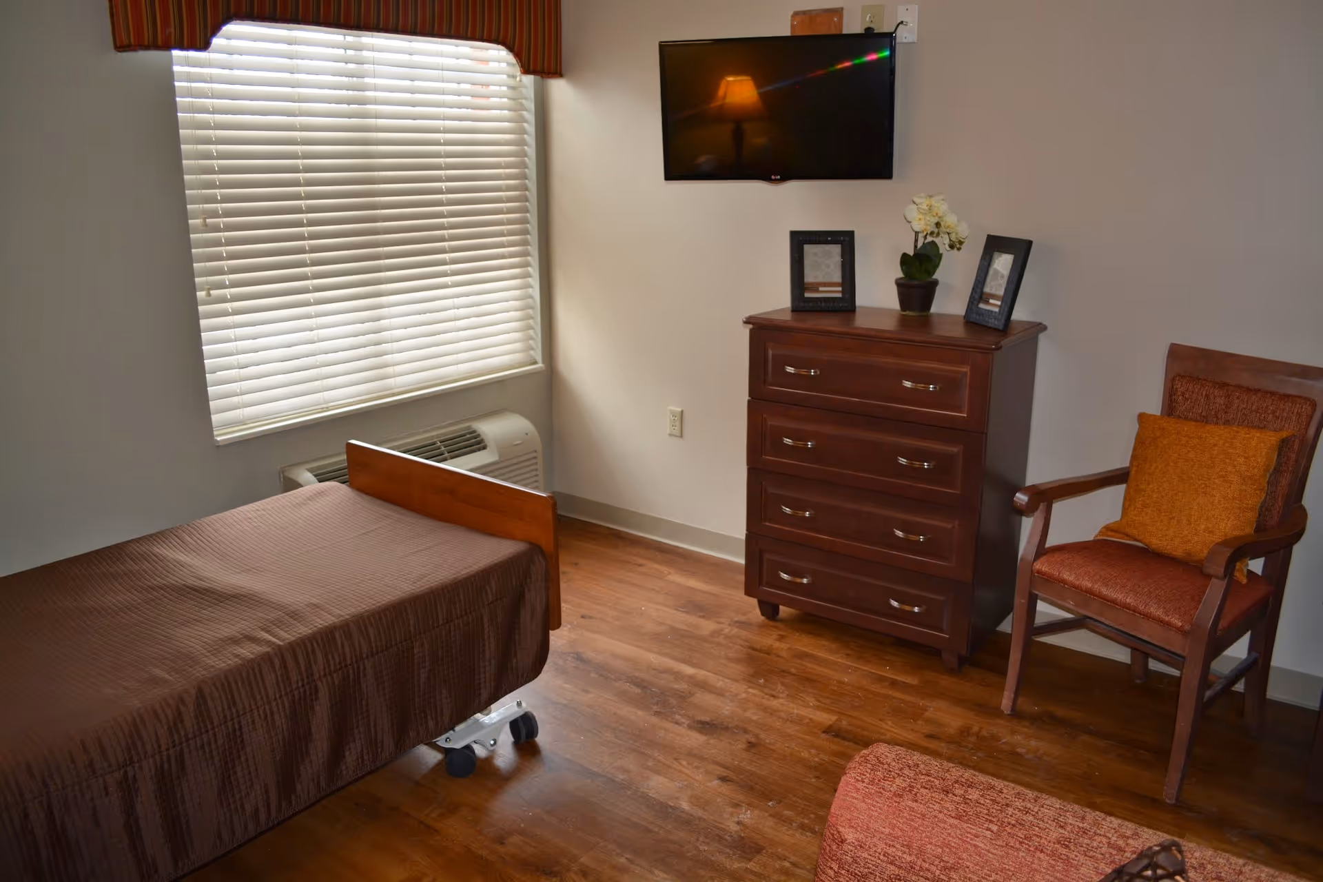 A small bedroom with a single bed covered in a brown bedspread, a wooden dresser with three drawers, a flat-screen TV mounted on the wall above the dresser, a wooden chair with an orange cushion, and a window with closed blinds and a striped valance. The floor is wooden and there is a small potted plant and two picture frames on the dresser.