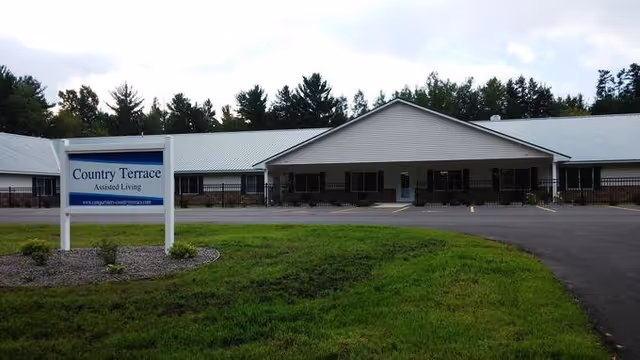 Exterior view of a single-story assisted living facility named Country Terrace, with a large sign in front displaying the facility's name and website. The building has a metal roof and is surrounded by a paved driveway and green lawn with trees in the background.