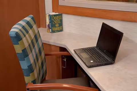 A desk with laptop, patterned chair, and a book against a wooden-framed wall.