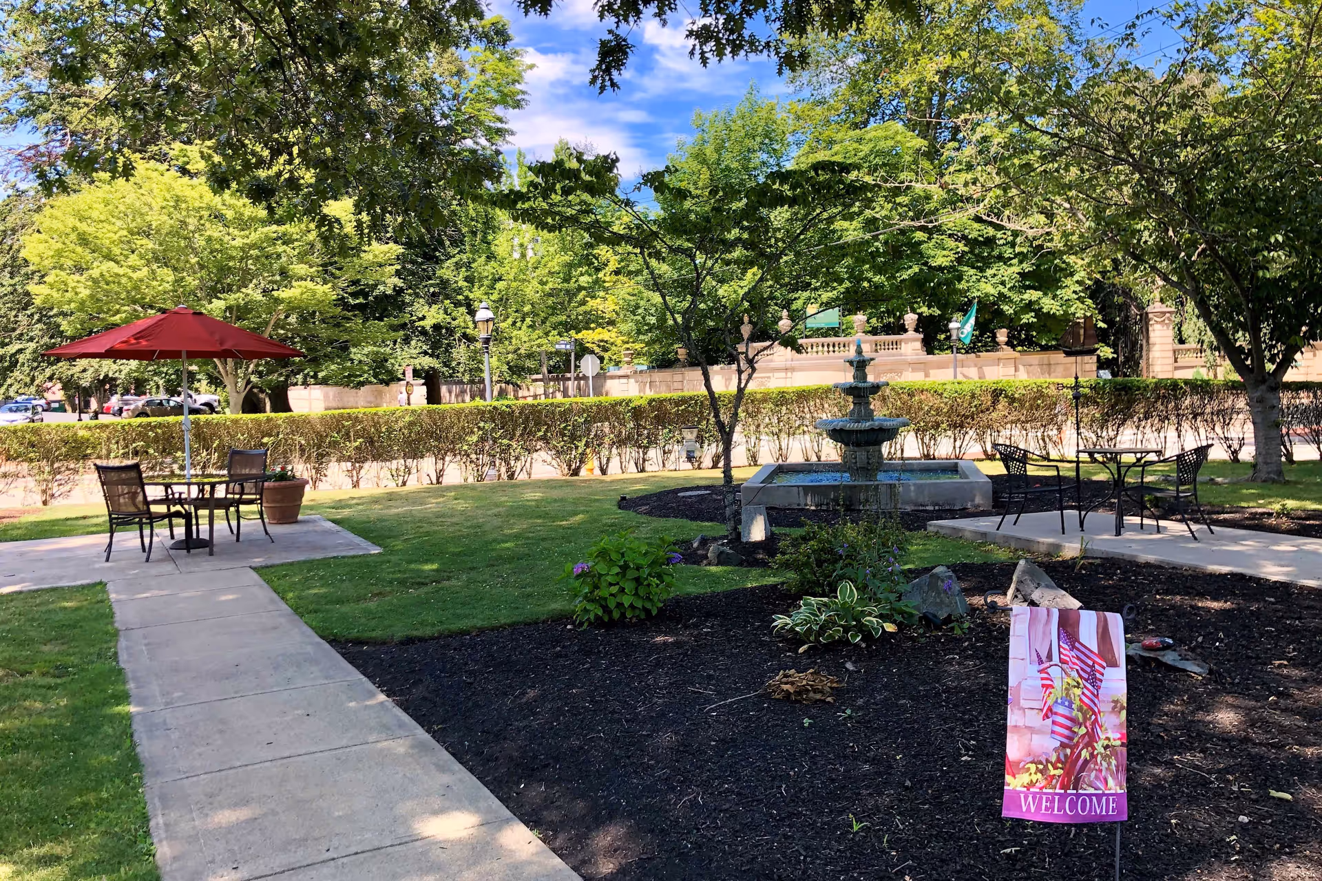 Outdoor garden area with green grass, trees, and shrubs. There are two seating areas with metal tables and chairs, one with a red umbrella. A stone fountain is visible in the background, and a small flag with the word 'WELCOME' is planted in the soil near the foreground. The sky is blue with some clouds.