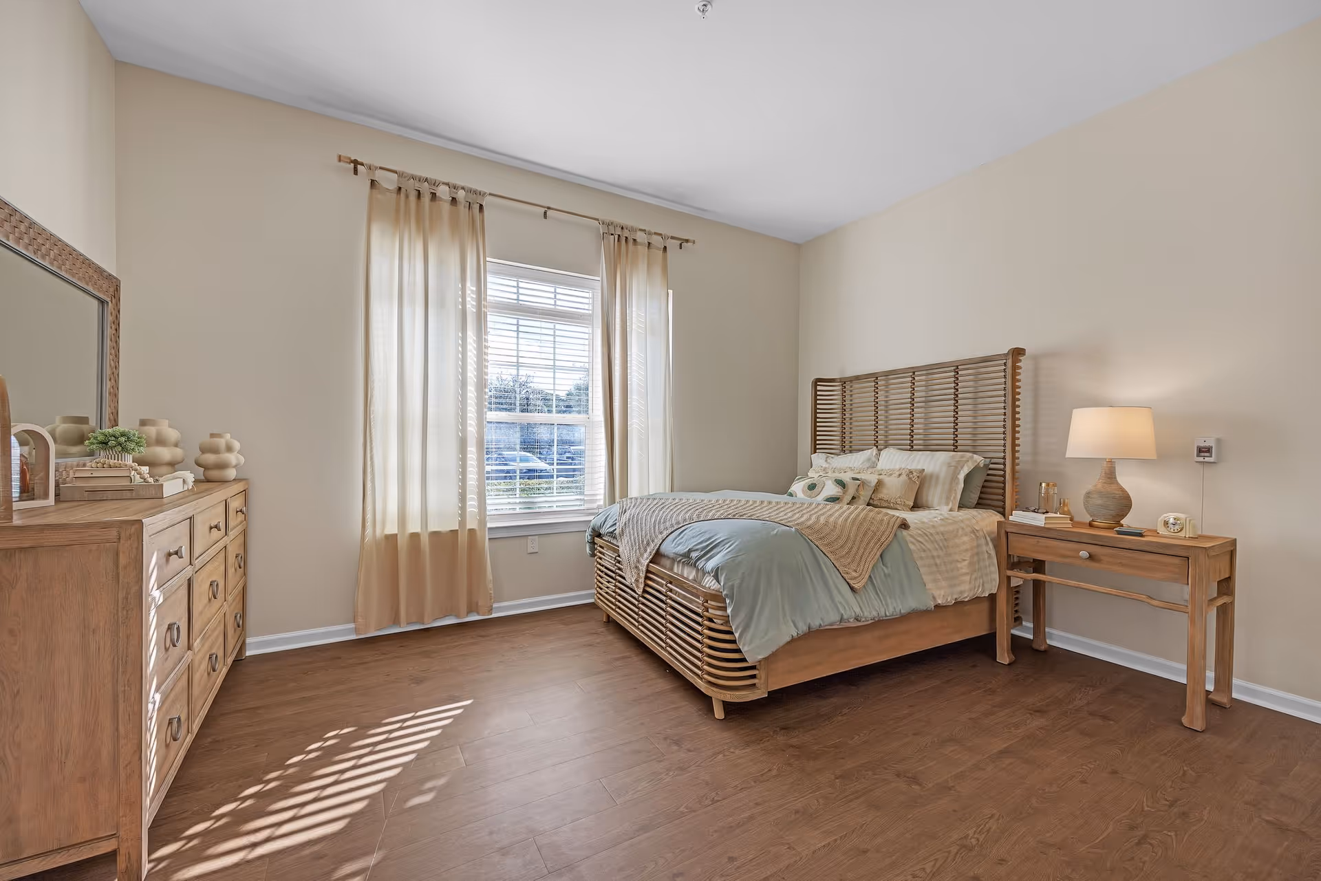 Sunlit bedroom with a wooden slatted bed and headboard, nightstand with lamp, dresser, and a window with beige curtains.